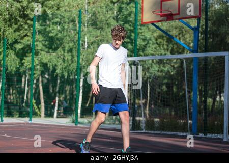 L'adolescente simpatico gioca a basket nel parco giochi della città. Un ragazzo tiene la palla di basket nelle mani esterne. Vita attiva, hobby, sport per bambini Foto Stock