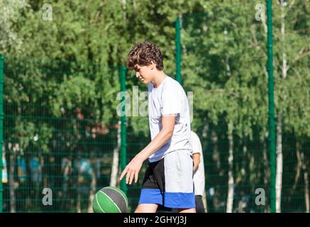 L'adolescente simpatico gioca a basket nel parco giochi della città. Un ragazzo tiene la palla di basket nelle mani esterne. Vita attiva, hobby, sport per bambini Foto Stock
