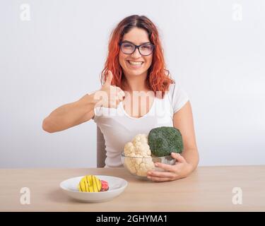 La donna caucasica preferisce cibo sano. Ragazza rossa sceglie tra broccoli e ciambelle su sfondo bianco. Foto Stock