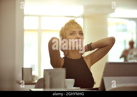 Ansiosa giovane donna preoccupata che medita su un problema con le sue mani aggrappate dietro la testa guardando verso l'alto al soffitto in un ristorante retroilluminato dalla s. Foto Stock