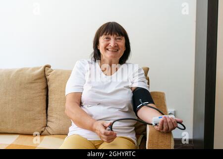 Donna adulta che misura la pressione sanguigna e il polso con un dispositivo elettronico. Donna matura a casa sul divano Foto Stock