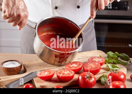 Non riconoscibile chef maschio di raccolto mescolando appetitosa zuppa di pomodoro in pentola mentre si prepara il pranzo in cucina a tavola di legno Foto Stock