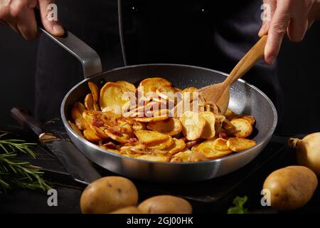 Crop faceless maschio cuoco preparazione gustosa patate arrosto in padella in cucina su sfondo scuro Foto Stock