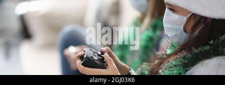 Due girlfriends sono seduti sul divano con i joystick di gioco in babbo natale cappelli e maschere mediche protettive Foto Stock