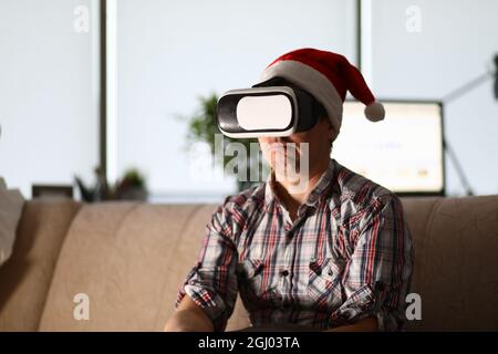 L'uomo in occhiali virtuali siede sul divano indossando il cappello di babbo natale Foto Stock