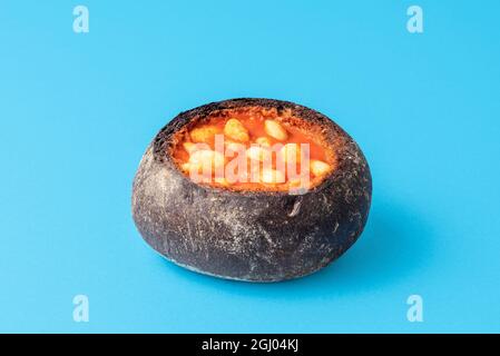 Fagioli cotti in un recipiente del pane su sfondo blu. Piatto tradizionale con fagioli al forno in crosta di pane minimalista su un tavolo. Foto Stock