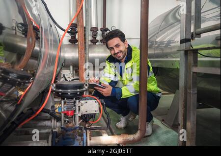 Happy uomo tecnico caucasico in uniforme checklist con appunti in pipeline d'acciaio e valvole in fabbrica di lavorazione Foto Stock