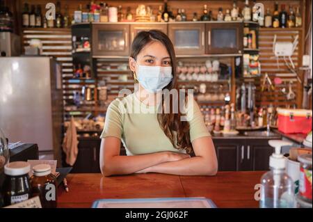 Attraente giovane imprenditore asiatico donna indossare maschera in piedi per accogliere i clienti al banco in legno nel ristorante Foto Stock