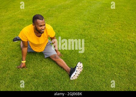 Sorridente maschio afroamericano in elegante abbigliamento sportivo seduto all'erba, cercando di fare una spaccatura, gambe stretching. L'uomo atletico sta lavorando, sta facendo Foto Stock