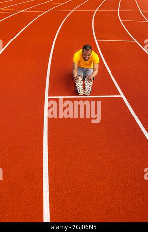 Atletica maschile in giallo abbigliamento sportivo seduto sul tracciato dello stadio, facendo un riscaldamento prima di correre, allungando le gambe. Uomo africano americano concentrato Foto Stock