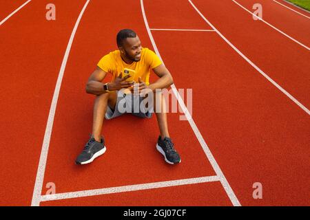 Felice atleta maschile attraente seduto sul circuito dello stadio, rilassarsi, fare una pausa dopo la corsa. Bell'uomo afroamericano che usa lo smartphone Foto Stock