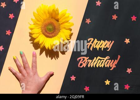 Testo Happy Birthday tagliato da carta arancione e rossa. La mano tiene il girasole naturale. Fiore in mano, piccoli elementi floreali su lavagna. Divisione diagonale Foto Stock