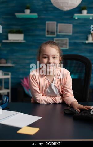 Ragazza piccola gioiosa sorridente seduta alla scrivania per il lavoro in linea della scuola su Internet lezione in aula. Bambino intelligente con materiali didattici penna notebook preparazione per la riunione di comunicazione Foto Stock