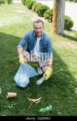 Uomo sorridente in guanti di gomma che tengono la bottiglia e la borsa dei rifiuti sul prato Foto Stock