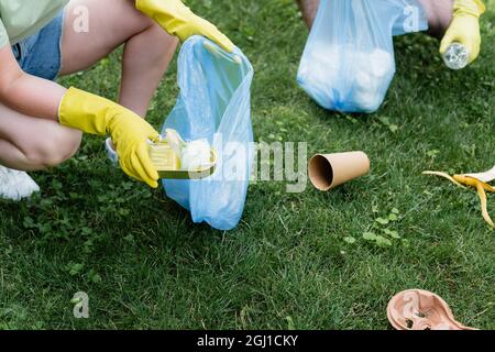 Vista ritagliata della donna in guanti di gomma che tengono lattina e sacco di rifiuti vicino a marito sfocato all'aperto Foto Stock