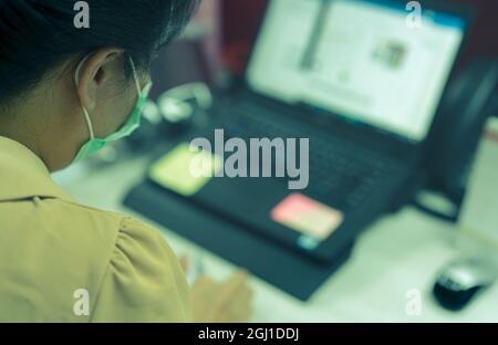 Vista posteriore di donna indossare maschera viso che lavora alla scrivania con computer portatile in ufficio. Donna asiatica che utilizza il computer in ufficio durante il lavoro da casa. Nuova normale Foto Stock