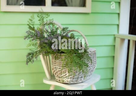 Cestino di vimini con bouquet di fiori selvatici estivi su sedia. Terrazza decorata con sedia e cestino con fiori. Elegante arredamento primaverile sulla veranda anteriore. Foto Stock