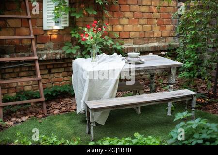 Terrazza esterna con muro in mattoni e piante, tavolo da pranzo e panca in legno. Giardino in estate con patio, mobili da giardino in legno ​in cortile Foto Stock