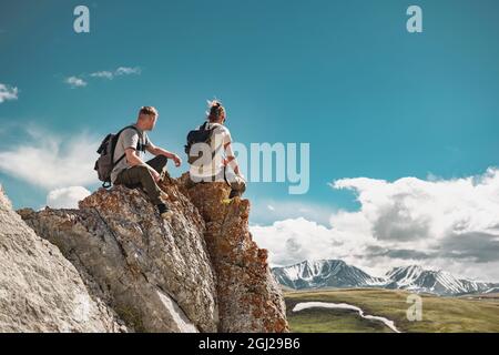 Due giovani escursionisti con piccoli zaini si rilassano sulla roccia grande o sulla cima della scogliera e si godono la vista della montagna Foto Stock