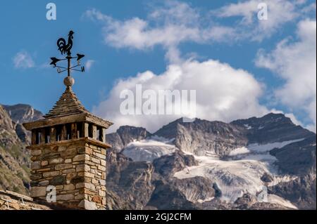 Una paletta meteorologica a forma di gallo in cima a un camino a Lillaz, Valle d'Aosta, Italia, con sullo sfondo il ghiacciaio del Gran Paradiso Foto Stock