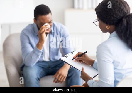 Concetto di salute mentale. Psicoterapeuta professionista che ha una seduta con il giovane uomo nero depresso piangendo in clinica, fuoco selettivo. Trattamento di Foto Stock