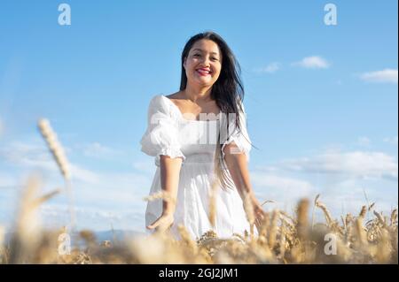 Giovane donna in abito bianco in piedi su campo di grano dorato in giorno di sole, toccando delicatamente il grano. Foto di alta qualità. Foto Stock