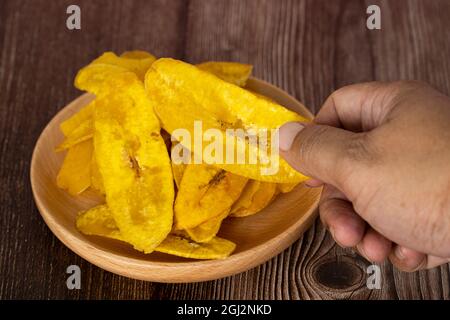 Mano che raccoglie un chip di banana o localmente conosciuto come Kerepek pisang da un piatto di legno. Sfondo sfocato Foto Stock