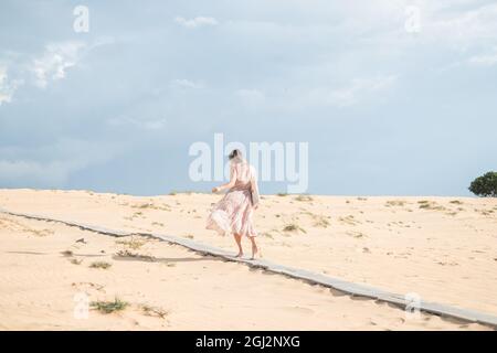 Bella vista della donna bionda caucasica in abito lungo a piedi nel deserto con sabbia bianca in giorno di sole durante le vacanze estive. Dune di sabbia. Foto Stock