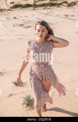 Ritratto di bella donna caucasica bionda in lungo abbigliamento casual a piedi nel deserto in giorno di sole durante le vacanze estive. Dune di sabbia. Foto Stock