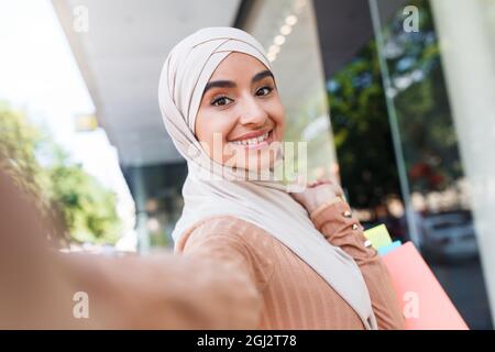 Allegro felice bella millennial arabo femmina buyer blogger in hijab con un sacco di borse rende selfie sul dispositivo vicino al centro commerciale, primo piano. Flusso di vita, moda Foto Stock