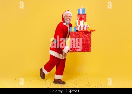 Ritratto a tutta lunghezza di uomo anziano con barba grigia in costume di babbo natale che tiene stack di presette natalizie, celebrando le vacanze di Natale. Poll Foto Stock