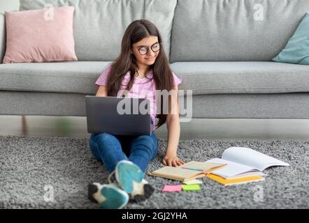 Focalizzata ragazza indiana teen seduta sul pavimento con laptop e materiali di studio, imparare online da casa, spazio libero. Adolescente intelligente pronto per r Foto Stock