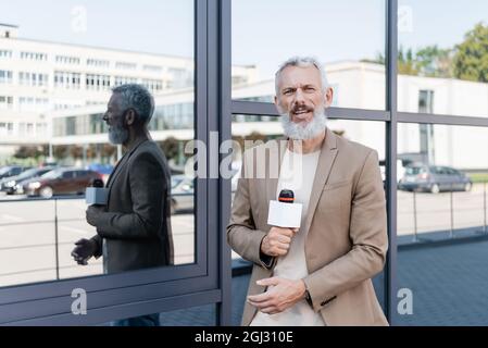 reporter bearded nel blazer che tiene il microfono e fa il reportage vicino all'edificio Foto Stock