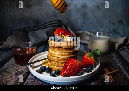 Una pila di pancake con miele versante, mirtillo e fragola e panna di frusta sul piatto su tavola di legno, delizioso dessert per la colazione Foto Stock