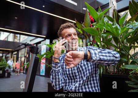 Uomo d'affari millenario che parla al telefono, uomo elegante sorridente controlla il tempo sul suo telefono. Ragazzo caucasico seduto accanto al suo scooter elettrico Foto Stock