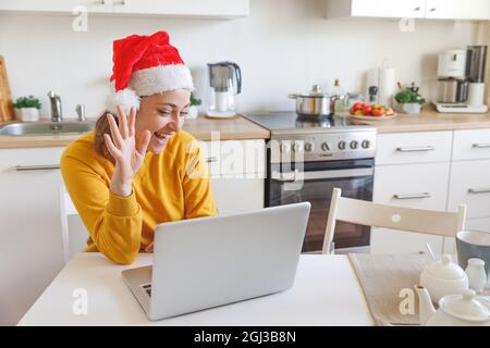 Ragazza sorridente che indossa il cappello di Santa che svela la mano videochiamata famiglia con la webcam. Donna con un portatile che ha una chat virtuale di riunione durante le vacanze seduti Foto Stock