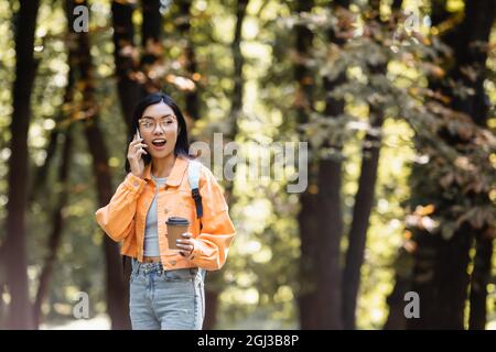 donna asiatica stupita con takeaway drink parlare sul telefono cellulare nel parco Foto Stock