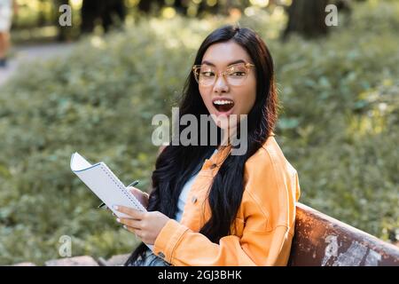 stupito studente asiatico con notebook guardare via mentre si siede sulla panca nel parco Foto Stock