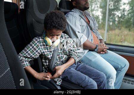 Ragazzo Afro americano serio con cuffie intorno al collo seduto in sedia di autobus e si piega prima della corsa Foto Stock