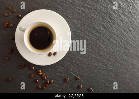 Tazza bianca di caffè sul piatto e fagioli sul tavolo dell'ardesia con spazio vuoto. Vista dall'alto. Composizione orizzontale. Foto Stock