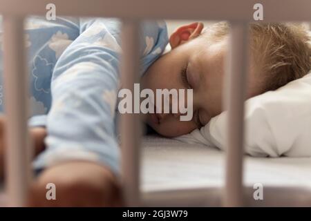 Carino piccolo allegro 2-3 anni prescolare bambino ragazzo dormire dolcemente in culla bianca durante il pranzo riposo tempo in blu pajama con cuscino a casa Foto Stock