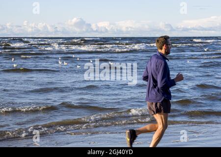 Esercitarsi all'esterno. Un jogger in t-shirt a manica lunga blu leggera, pantaloncini e sneakers running si allena alla maratona, attraversando la spiaggia del Mar Baltico, sul vento nuvoloso di mattina presto Foto Stock