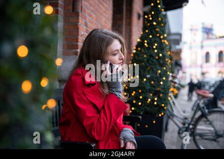 La ragazza fa una chiamata telefonica seduta su una panca vicino agli alberi di Natale decorati con le luci. Le luci di Natale bokeh con fuoco sulla donna - Natale Foto Stock