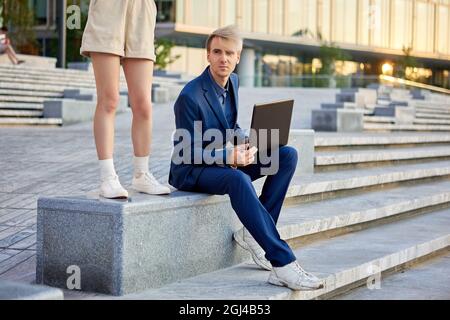 Gambe femminili in pantaloncini e uomo in tuta da lavoro con computer portatile vicino all'edificio dell'ufficio. Foto Stock