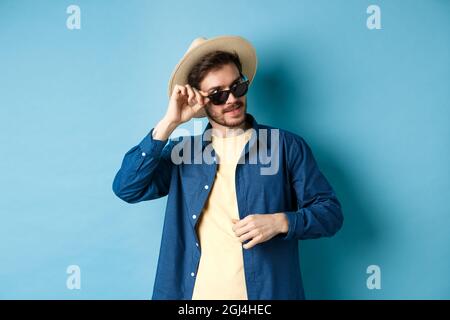 Bel turista in occhiali da sole e cappello di paglia guardando da parte, sorridente soddisfatto, esplorando avvistare in vacanza, sfondo blu Foto Stock