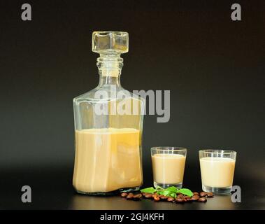 Liquore di caffè cremoso, bottiglia, due bicchieri, una manciata di fagioli tostati e foglie di menta su sfondo nero. Primo piano. Foto Stock