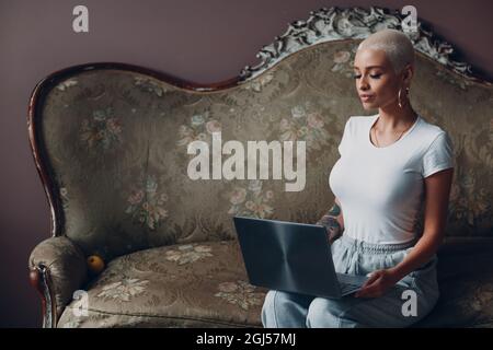 Giovane donna millenaria con ritratto di capelli biondi corti seduti con un computer portatile su un divano vintage in casa d'ufficio Foto Stock