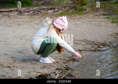 Una bambina siede sulle rive del fiume e tocca la superficie dell'acqua fredda con le proprie mani. La bambina gioca sulla riva del fiume. A lei Foto Stock