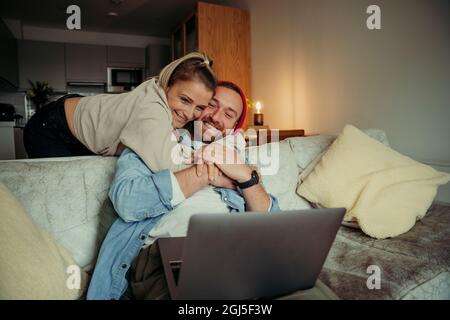 Coppia caucasica felice seduta sul divano che mostra l'amore mentre si abbraccia e coccola durante le videochiamate utilizzando il portatile Foto Stock