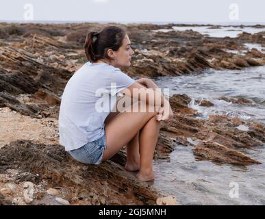 Ragazza teenage pensierosa seduta sulla scogliera vicino al mare con le gambe in acqua al tramonto e guardando dritto, indossando t-shirt blu e jeans shorts, laterale vie Foto Stock